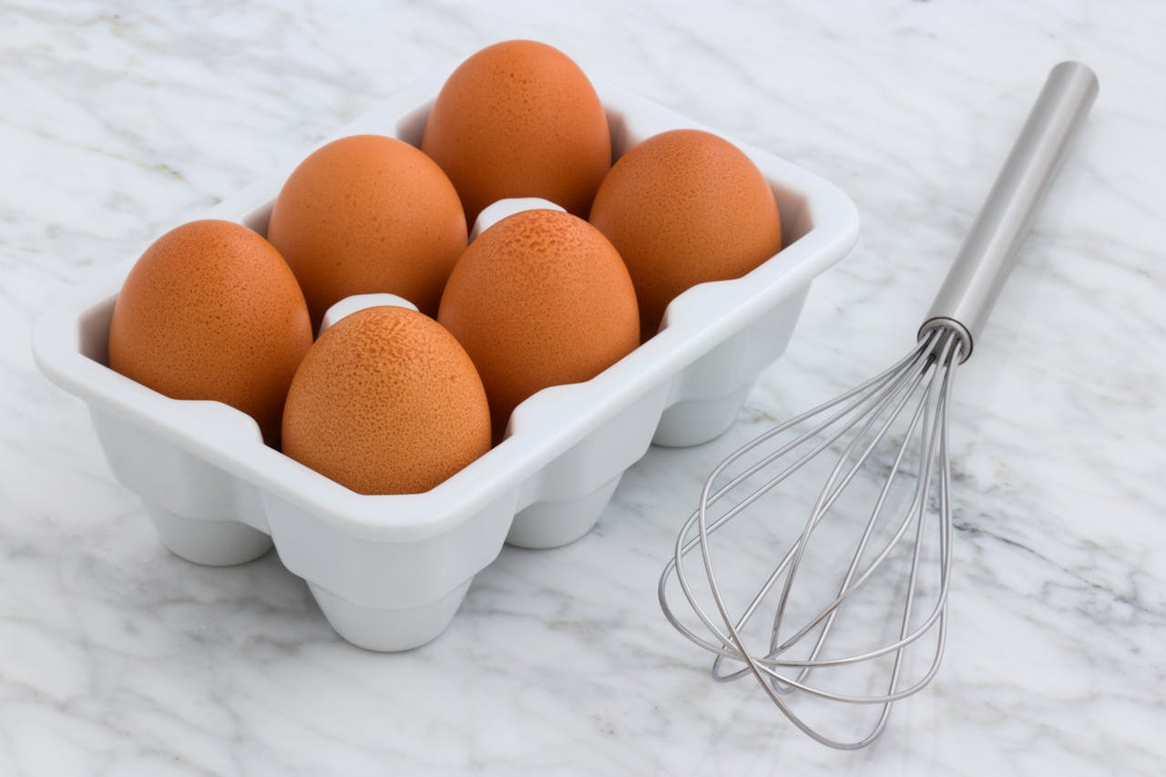 A tray of farm-fresh brown eggs alongside a whisk on a marble countertop, perfect for cooking inspiration.